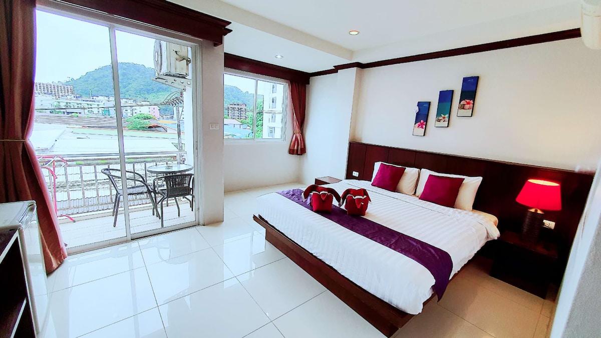 A bright room featuring a spacious bed adorned with two decorative red pillows. A window allows natural light to brighten the space, offering a view of the surrounding area. A small balcony with two chairs is visible, enhancing the room's inviting layout.