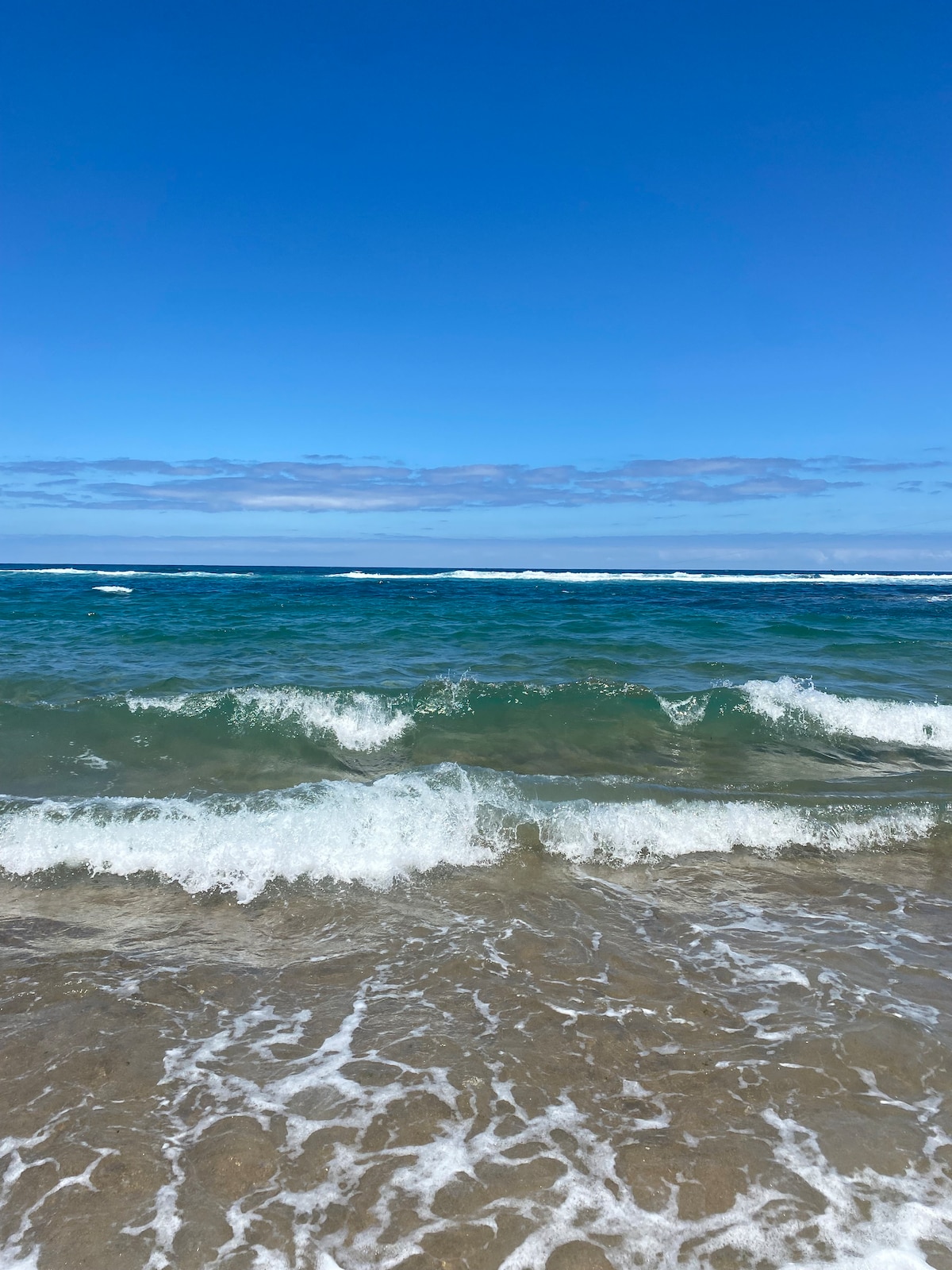 Gentle waves lapping against a sandy beach are visible, with the ocean reflecting shades of turquoise and blue. A clear sky stretches overhead, dotted with wisps of clouds, enhancing the serene coastal atmosphere.
