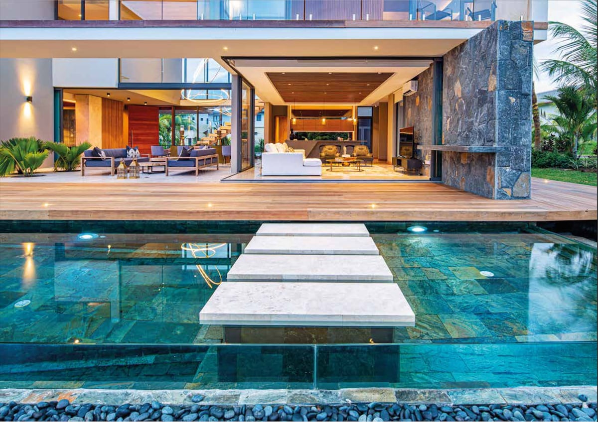 A luxurious outdoor area showcases a spacious terrace with modern seating arrangements and a sleek design. An infinity lap pool features a unique stepping stone pathway leading to a covered patio, highlighting the seamless connection between indoor and outdoor living spaces.