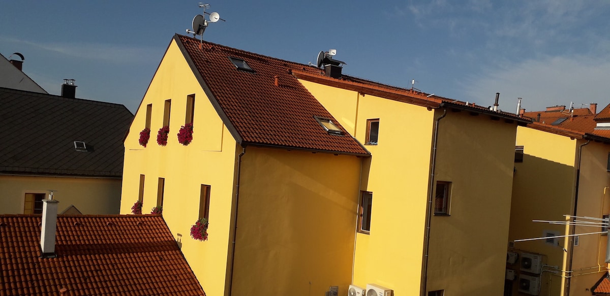 A vivid yellow building is showcased, featuring multiple windows and flower boxes that add a touch of color. The roof is lined with red tiles, and several air conditioning units are visible along the exterior. A clear blue sky adds to the backdrop.