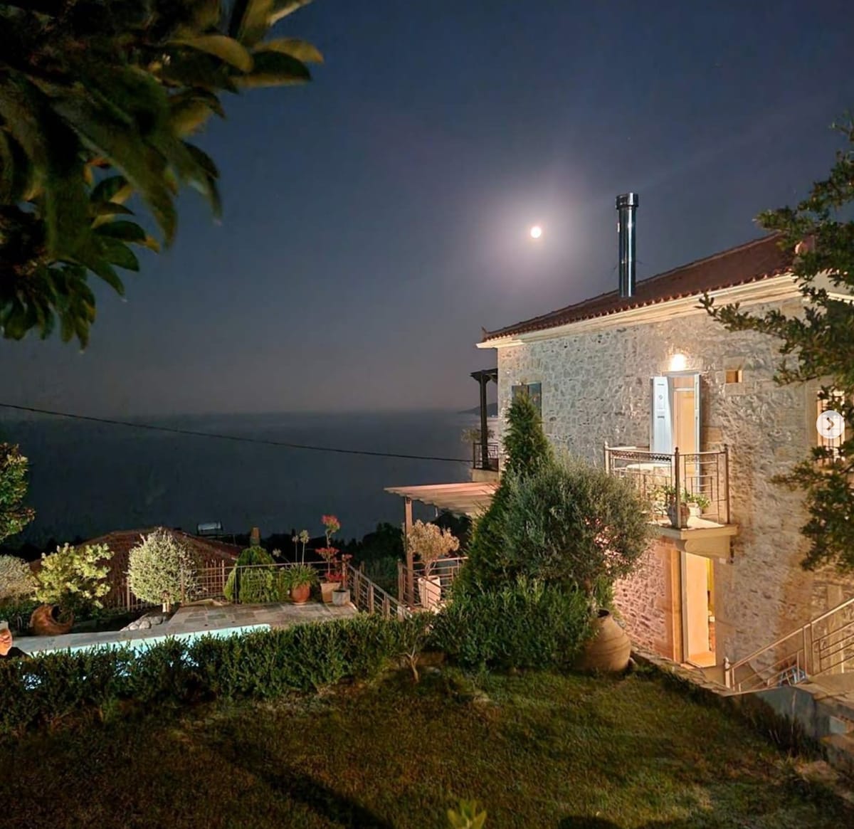 The stone house is seen at night, bathed in the glow of moonlight. A lush garden surrounds the property, highlighting various potted plants. The swimming pool reflects the soft light, and a chimney can be noted rising from the rooftop, offering a glimpse of a warm gathering space.