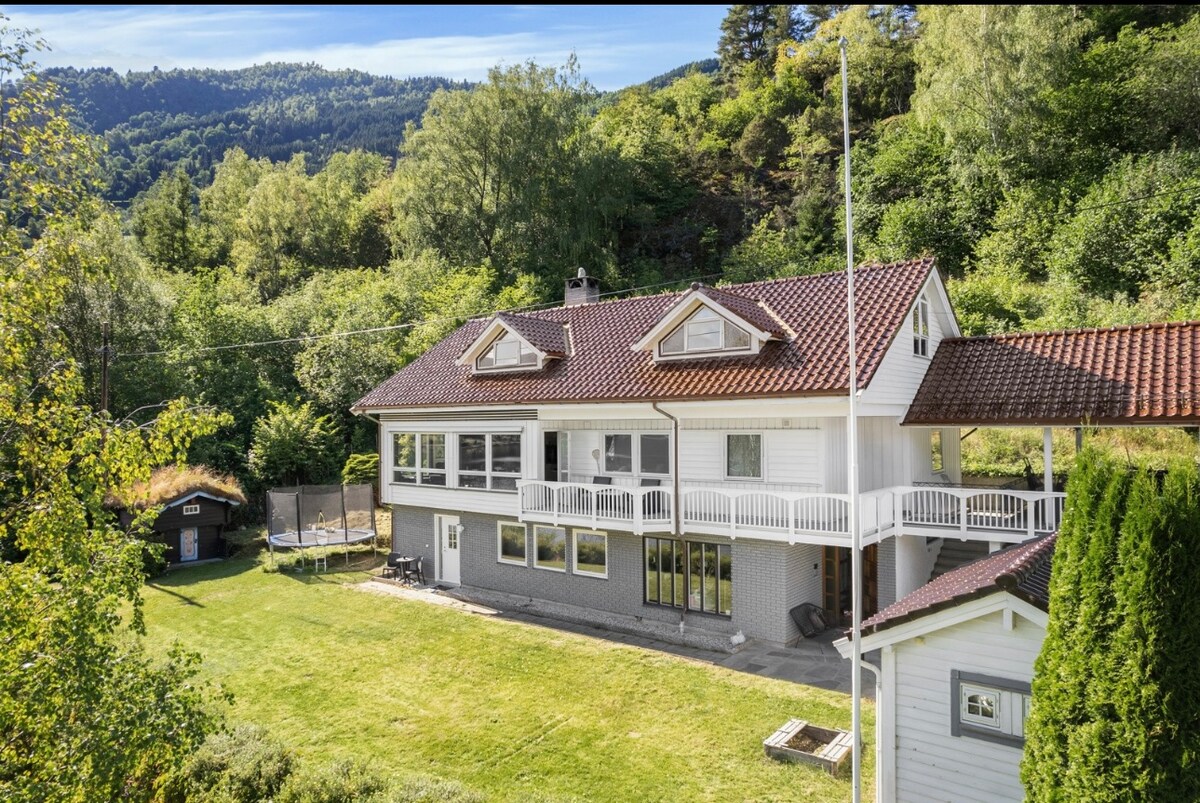 A spacious two-story house is presented, showcasing a large outdoor area surrounded by greenery. The building features multiple balconies with a red-tiled roof, large windows for natural light, and a well-maintained lawn. Scenic hills can be seen in the background.