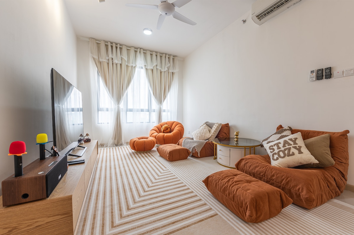 An inviting living area features plush seating in warm orange tones and a neutral striped rug. Large windows are adorned with light, flowing curtains, allowing natural light to fill the space. A simple entertainment unit holds a television and decorative accents, creating a cozy atmosphere.