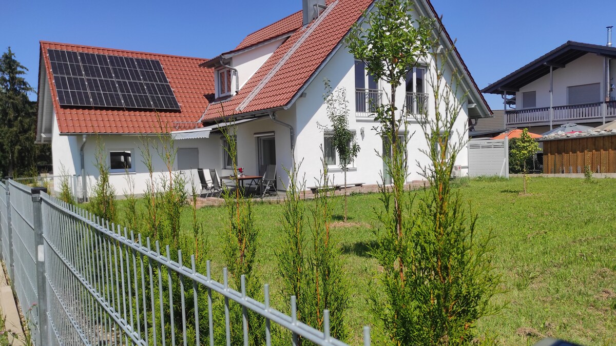 A modern two-story house is set against a clear blue sky, featuring a red tiled roof and solar panels. A well-maintained lawn is bordered by a metal fence with young trees planted throughout, creating an inviting outdoor space.