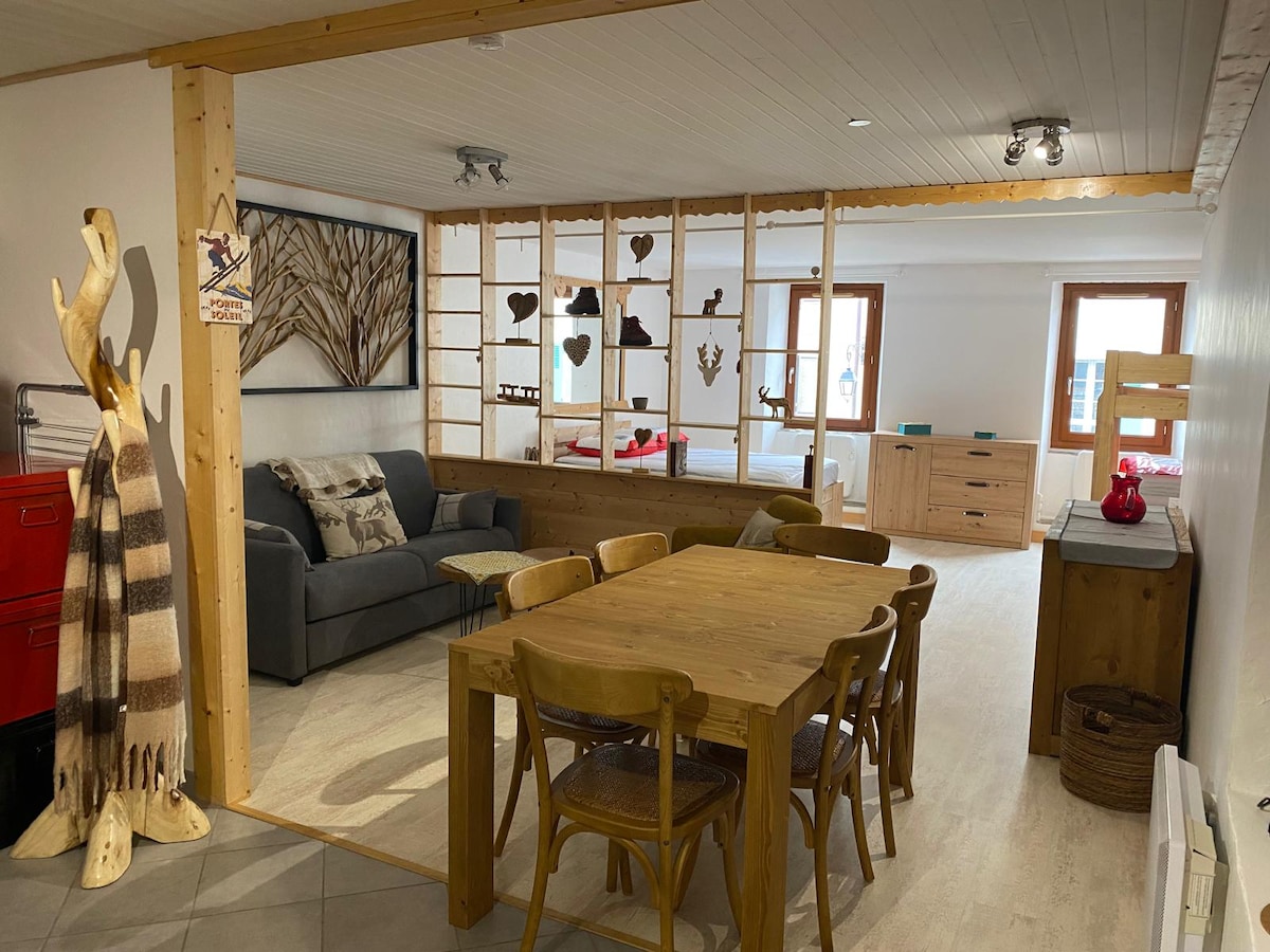 A spacious living area features a wooden dining table surrounded by six chairs. A comfortable sofa sits in the corner, complemented by a decorative wall with a mix of rustic and artistic elements. Natural light floods the open layout, highlighting the warm wooden accents.