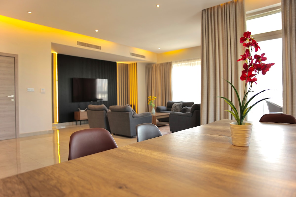 The open living area features modern seating arrangements with soft gray sofas positioned around a central coffee table. A large flat-screen TV is mounted on the wall, complemented by warm lighting from recessed fixtures. A dining table is set nearby, adorned with a potted plant.