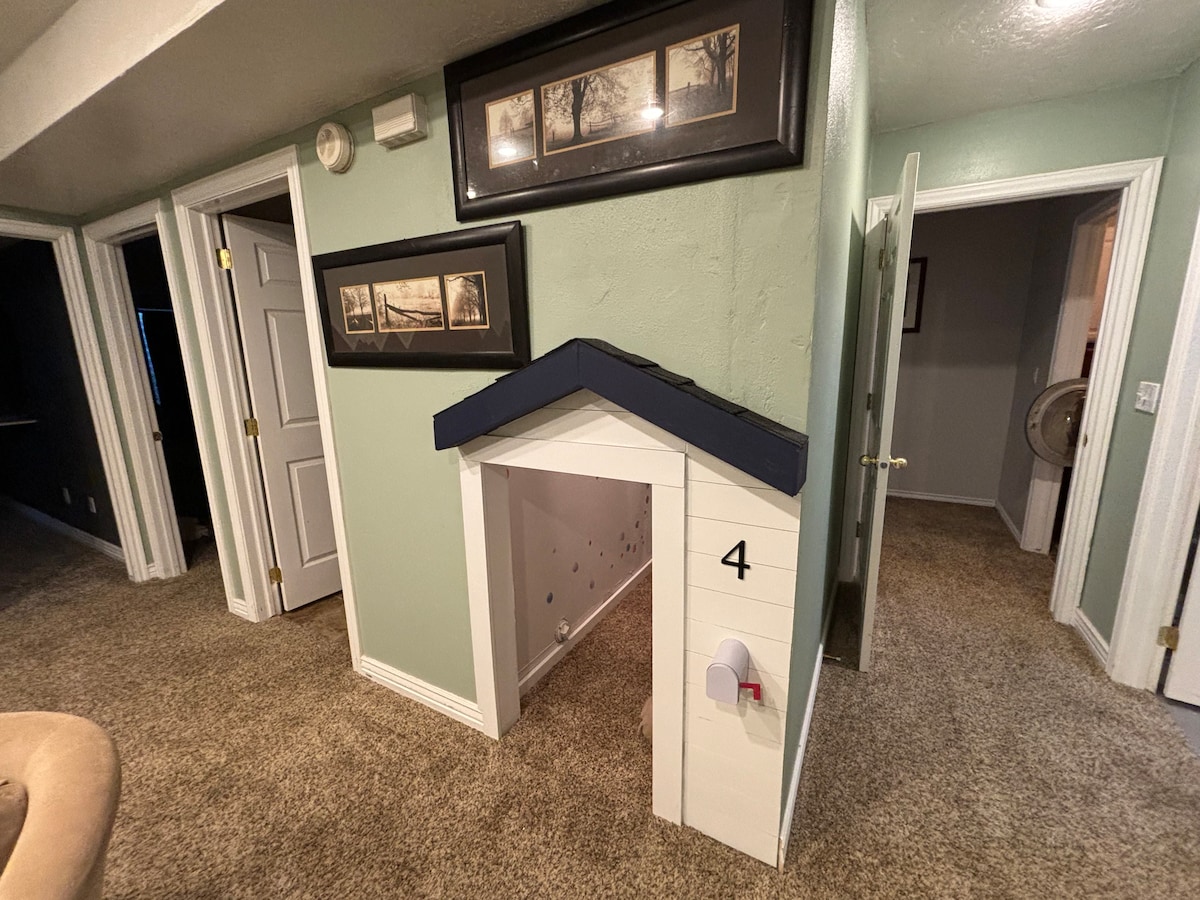 A charming playhouse is situated within a hallway, featuring a triangular roof and a door marked with the number 4. The walls are painted in soft green, and the carpeted floor offers a comfortable feel. Nearby, two doorways lead to different rooms.