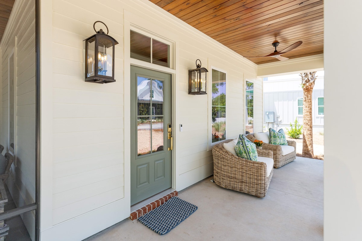 The entrance features a green door with glass panels, flanked by two lantern-style light fixtures. Comfortable seating is provided on a covered porch, with decorative pillows enhancing the inviting space. Natural light enters through large windows, illuminating the area and showcasing the wooden ceiling.