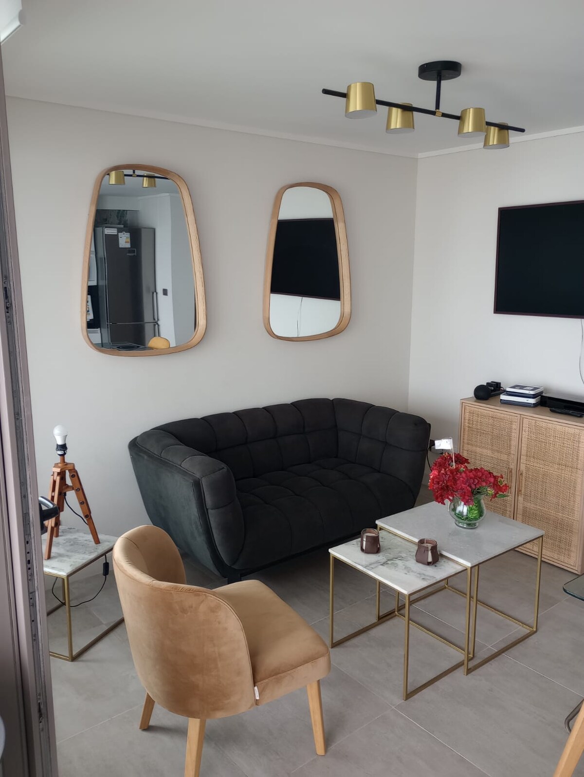 A modern living area features a dark, tufted sofa against a neutral wall. Two oval mirrors are positioned above the sofa, reflecting light. A wooden coffee table sits between the sofa and a gold-accented chair, complemented by decorative flowers and a wall-mounted television.