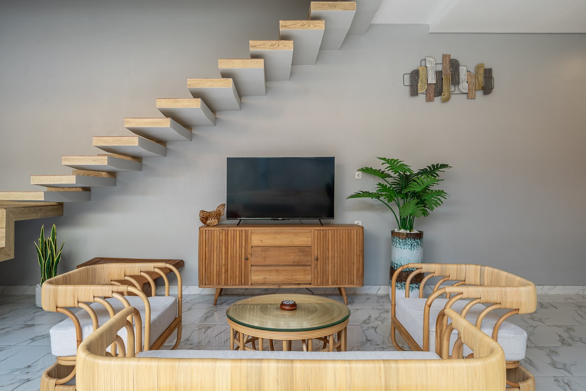 A modern living area features a minimalist design with light wood furniture and a glass coffee table. A flat-screen television is positioned on a stylish wooden console. Decorative plants and wall art add subtle accents to the space, complemented by neutral wall tones.