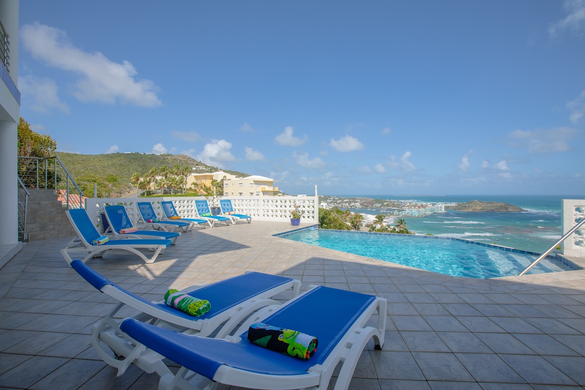 Lounge chairs are arranged by the infinity pool, providing views of the ocean and surrounding landscape. A tiled deck complements the outdoor space, while colorful beach towels rest on the chairs. The clear sky enhances the inviting atmosphere of this area.