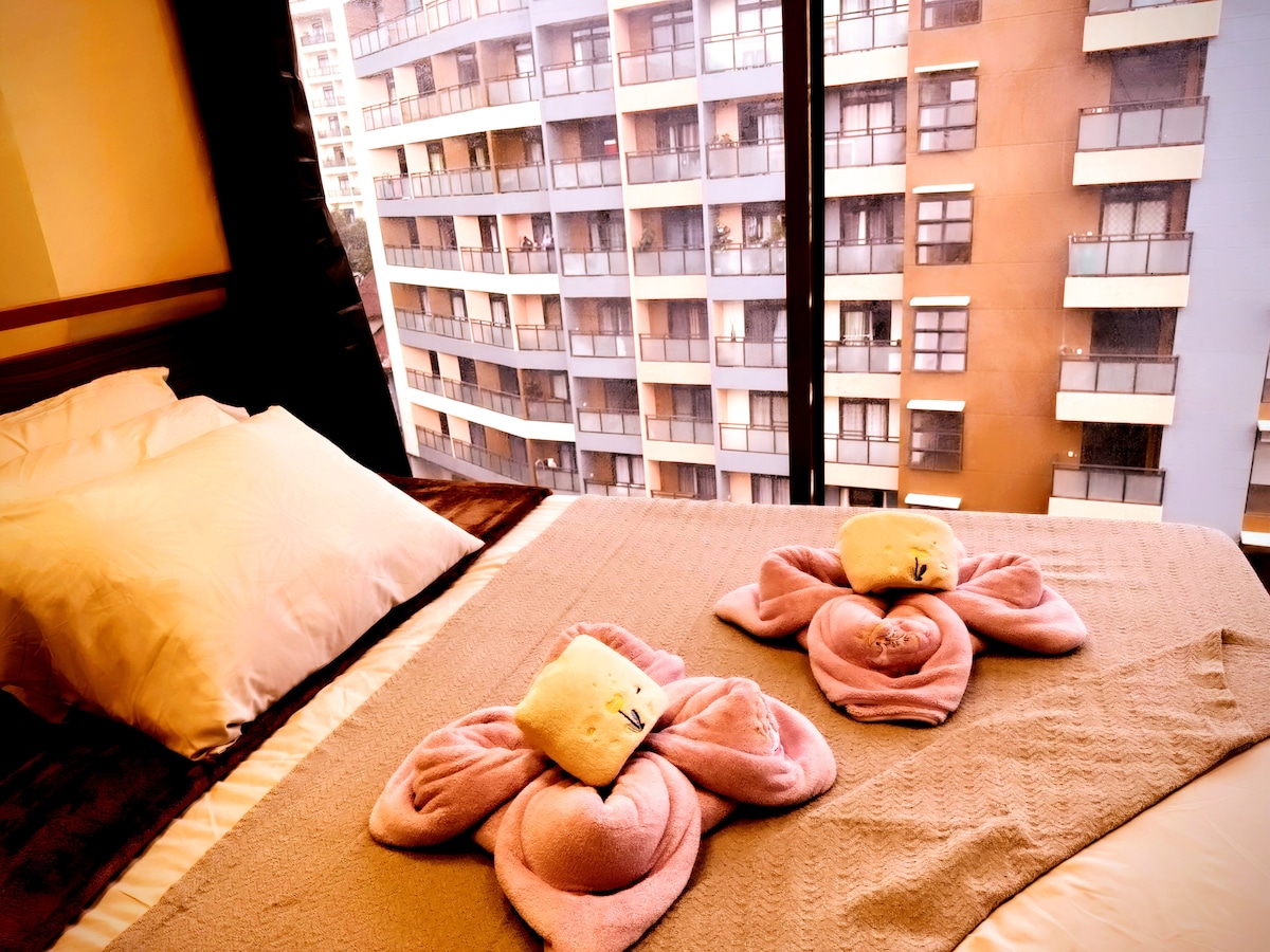 A spacious bed is adorned with soft linens, featuring unique towel arrangements resembling animals. Large windows offer a view of neighboring buildings, allowing natural light to fill the room.