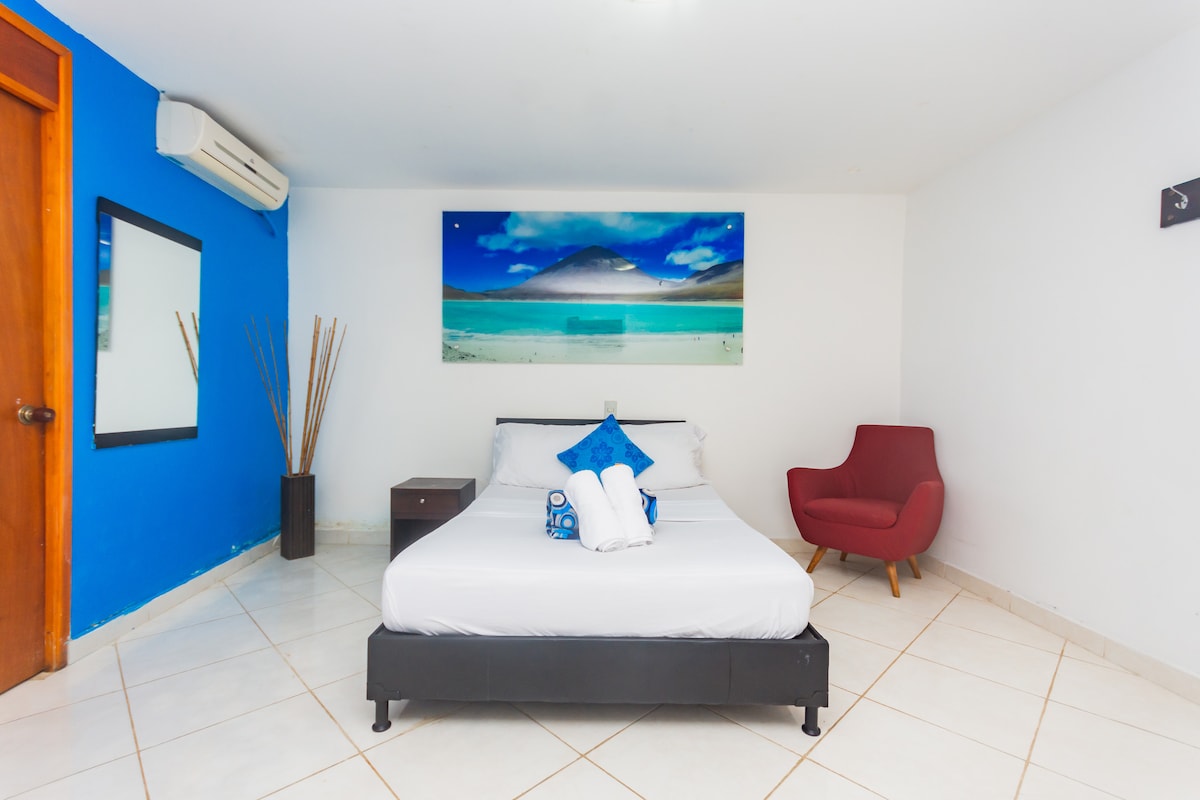 A spacious guest room features white tiled flooring and a large bed with neatly arranged towels. A vibrant blue accent wall complements a piece of landscape artwork. A red chair is positioned next to a bedside table, with a mirror reflecting the room's natural light.