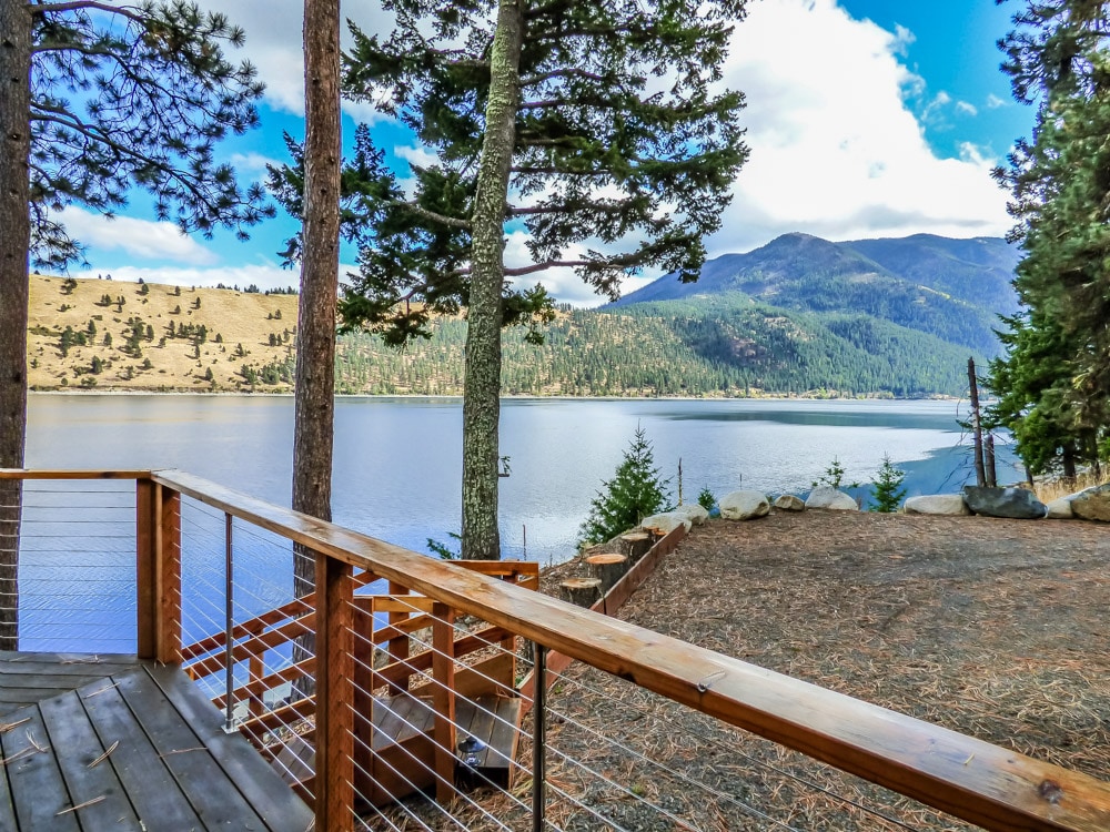 A wooden deck extends towards a serene view of Wallowa Lake, framed by tall trees. The landscape features gentle hills in the background, showcasing a blend of greenery and earthy tones under a partly cloudy sky.