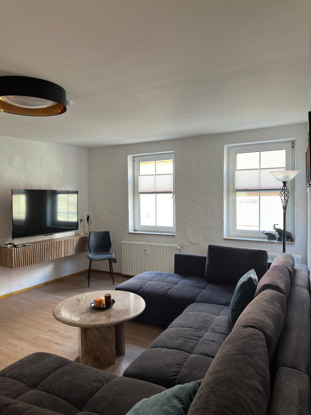 A comfortable living area features a dark sectional sofa and a marble coffee table. Two windows allow natural light, illuminating the space. A wall-mounted television is positioned across from the seating, and a chair sits nearby, offering additional seating options.
