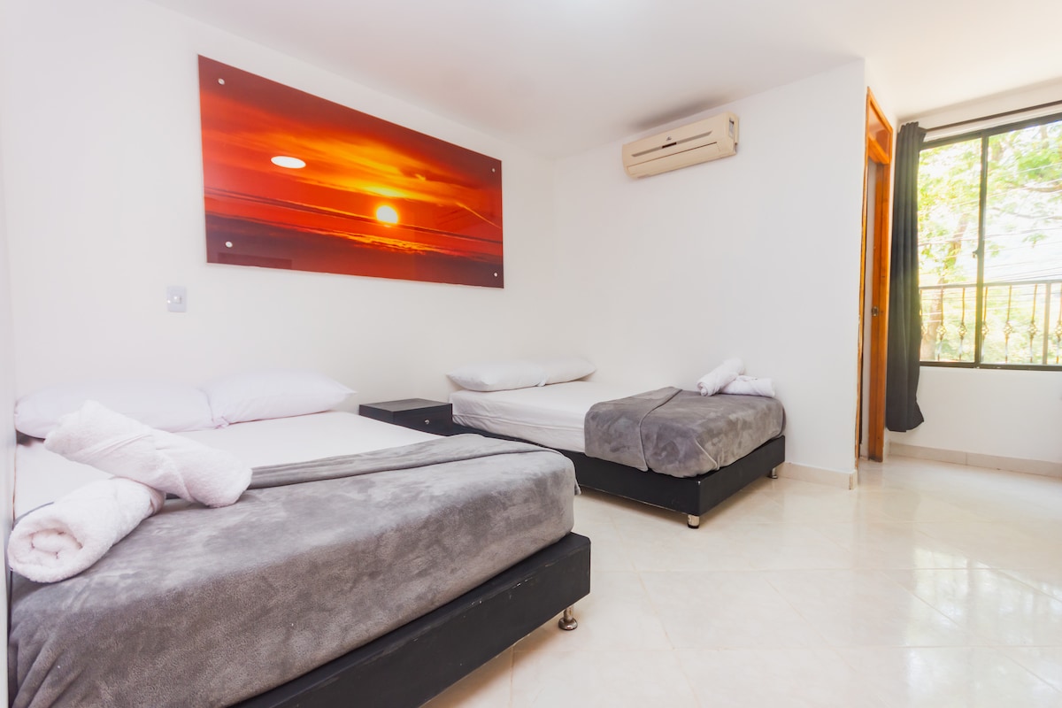 A bright guest room features two spacious beds covered with soft gray blankets. A large wall art piece displays a sunset over the ocean. Natural light washes over the room through a window with light curtains, and a small closet is visible near the entrance.