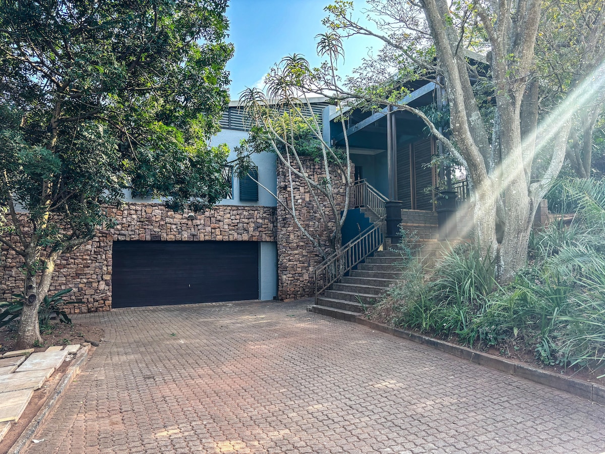 The home features a natural stone façade with a covered entrance, surrounded by lush greenery. A neatly paved driveway leads to the garage, while stone steps ascend to the main entry, inviting guests into the property.