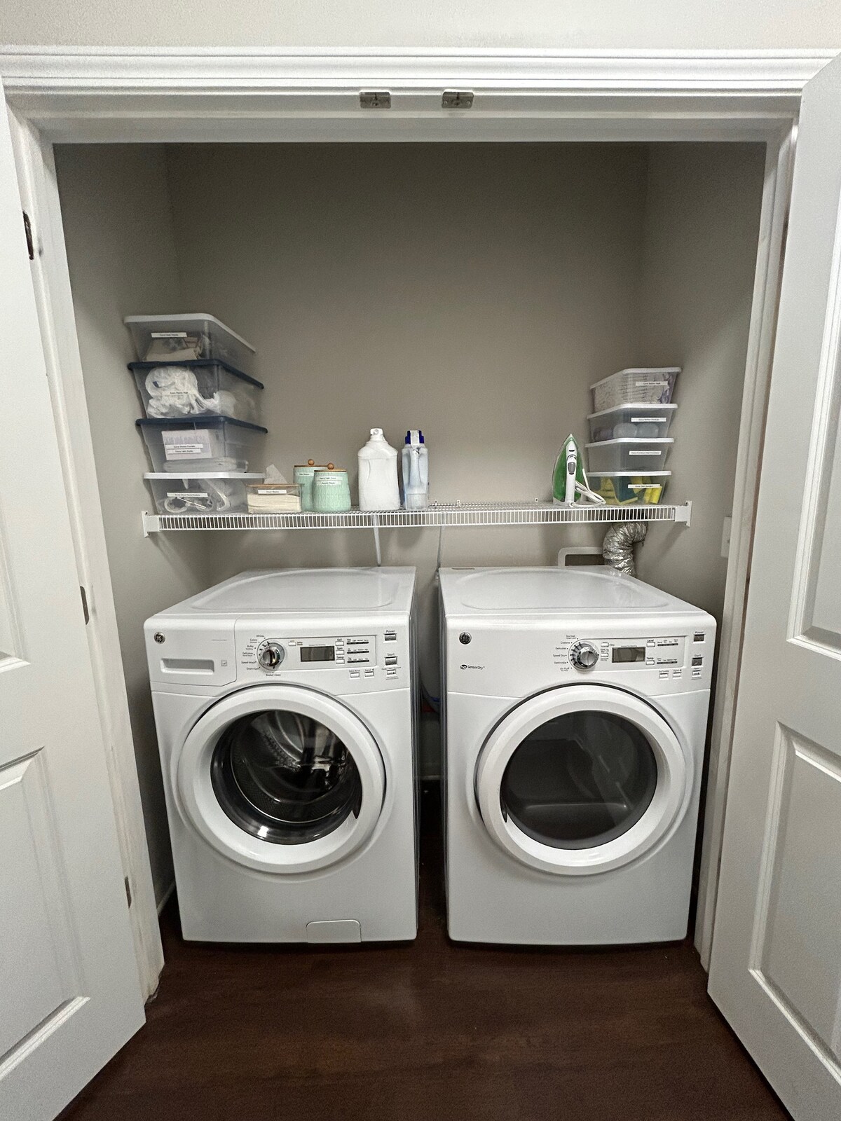 A dedicated laundry area features a stacked washer and dryer, both in white finishes. Above, a shelf holds several containers and cleaning supplies, providing organized storage. The closet is designed with light walls and a dark wood floor for a clean and functional space.