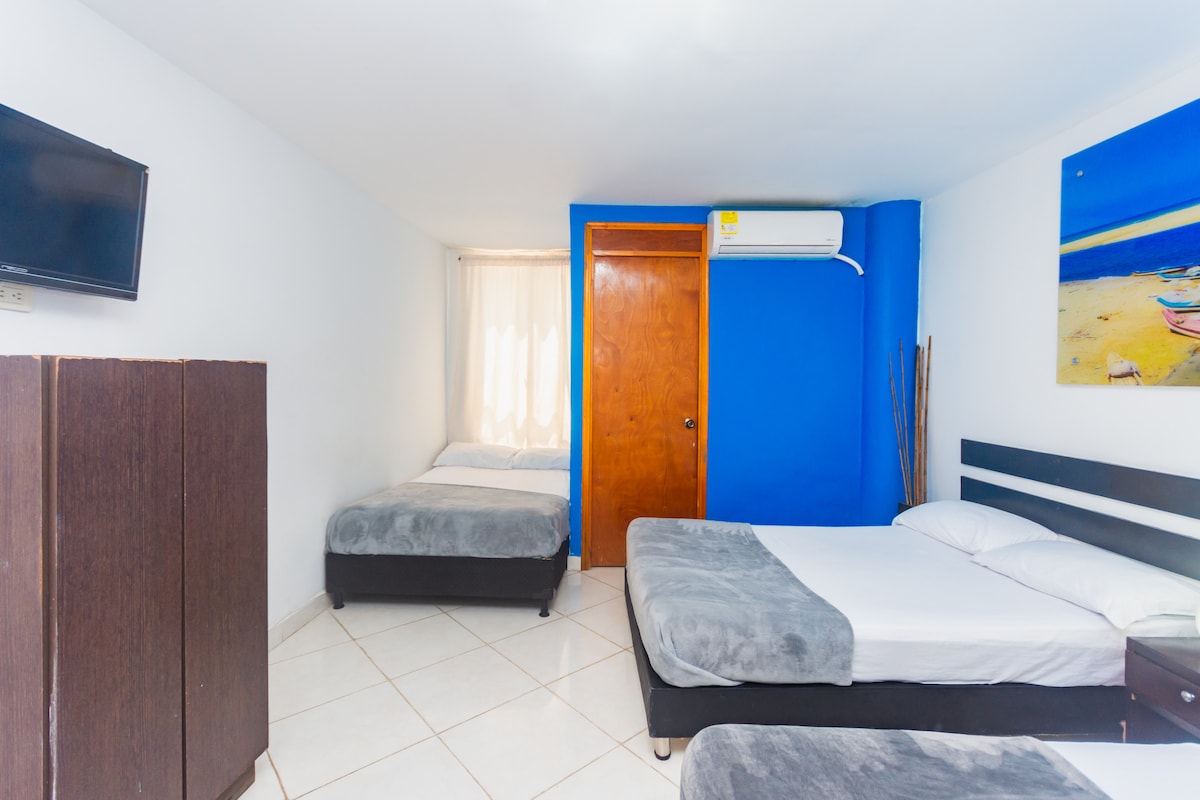 A comfortable guest room is presented with two spacious beds and a television mounted on the wall. Light filters through sheer curtains, illuminating the vibrant blue wall. A wooden door provides access to the room, while a colorful artwork adds a touch of visual interest.