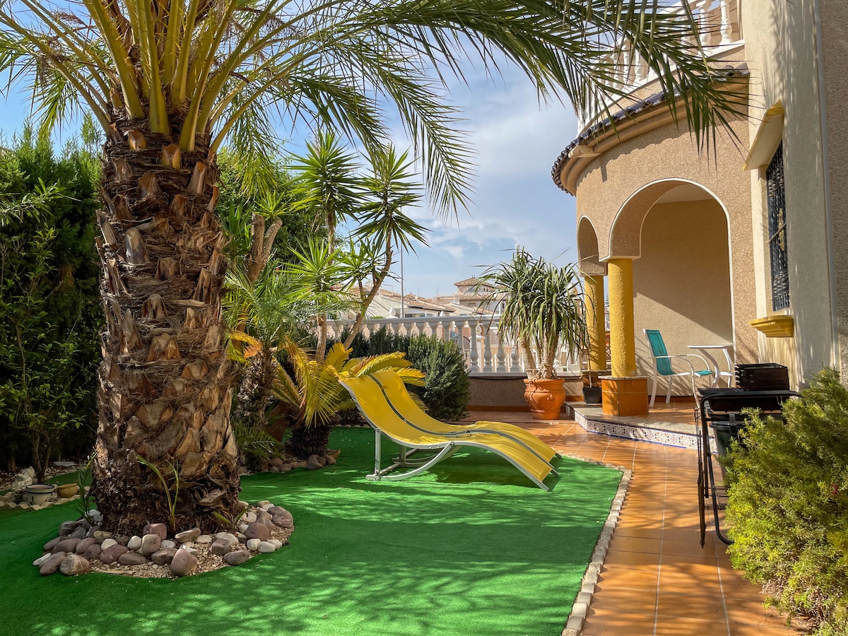 A green garden space is visible, featuring a large palm tree and various shrubs. A yellow lounge chair is positioned on the artificial grass, with decorative rocks surrounding it. A patio area is shown, framed by the rounded architecture of the building.