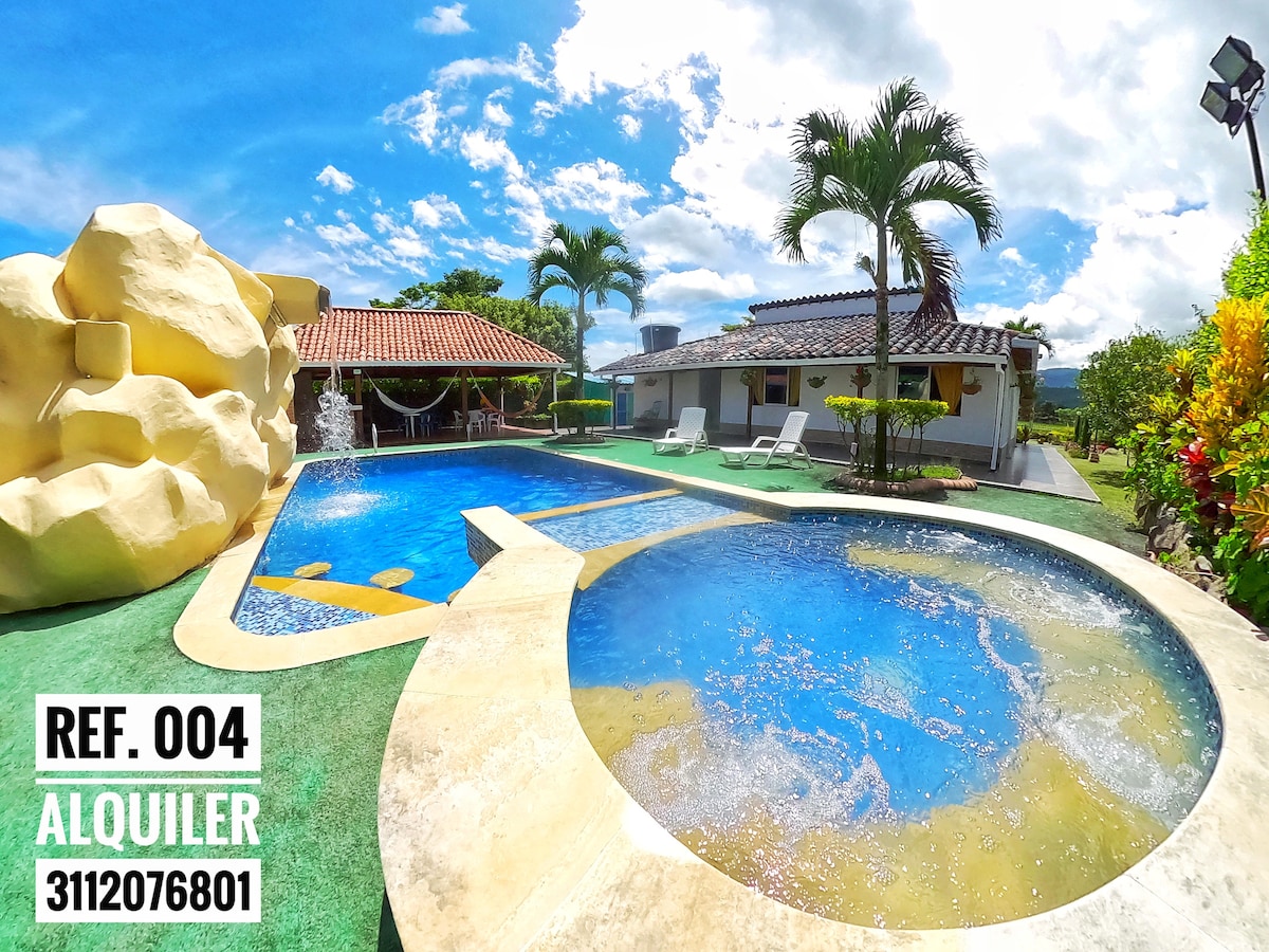 A vibrant outdoor area features a large swimming pool with a built-in jacuzzi. Surrounded by lush greenery, lounge chairs invite relaxation. The well-maintained stonework and colorful landscaping create an appealing environment, while a charming house with a clay tile roof is visible in the background.