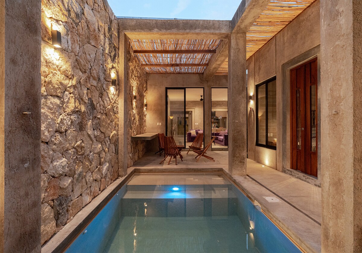 An inviting outdoor space features a small pool surrounded by stone walls and a thatched roof overhead. Two rustic chairs are positioned nearby, and large glass doors lead to an interior area. Soft lighting enhances the serene atmosphere of the space.