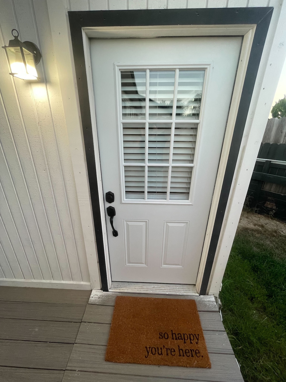 A welcoming entryway is shown, featuring a white door with decorative window panes and a black handle. A soft brown doormat with the phrase 'so happy you're here' adds a personal touch, while a light fixture on the wall provides visibility at night.