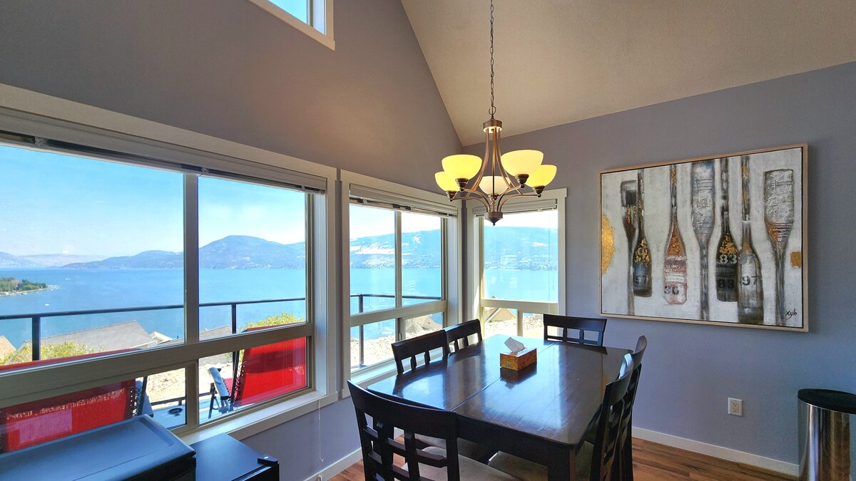 A dining area features a wooden table surrounded by six chairs, positioned near large windows offering expansive lake views. Natural light illuminates the space, highlighting a wall art piece depicting wine bottles. The design combines functionality and scenic beauty.