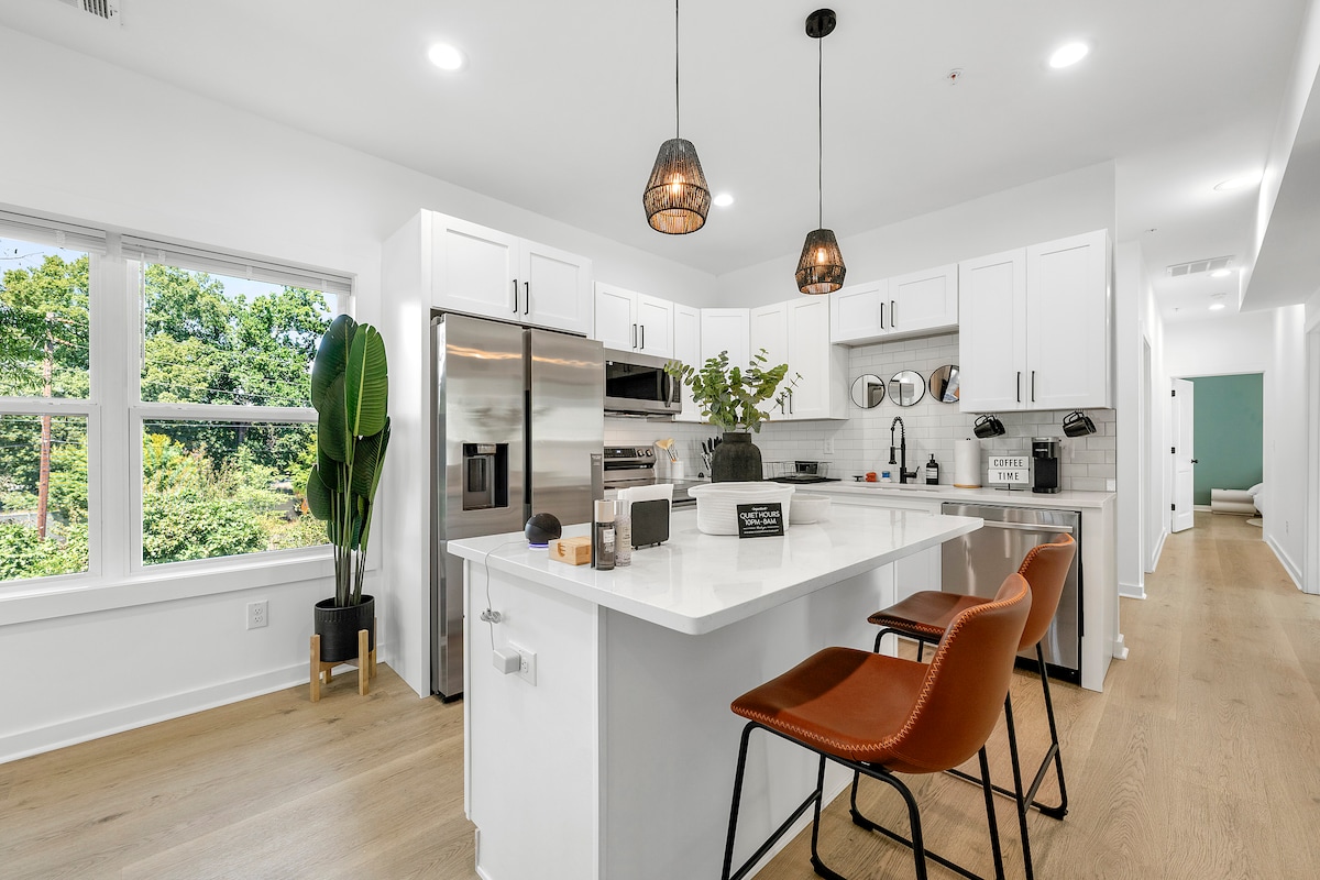A modern kitchen space is showcased, featuring bright white cabinetry and stainless steel appliances. A sleek kitchen island is adorned with stylish bar stools, while large windows reveal views of greenery outside. Decorative plants add a touch of nature to the inviting atmosphere.