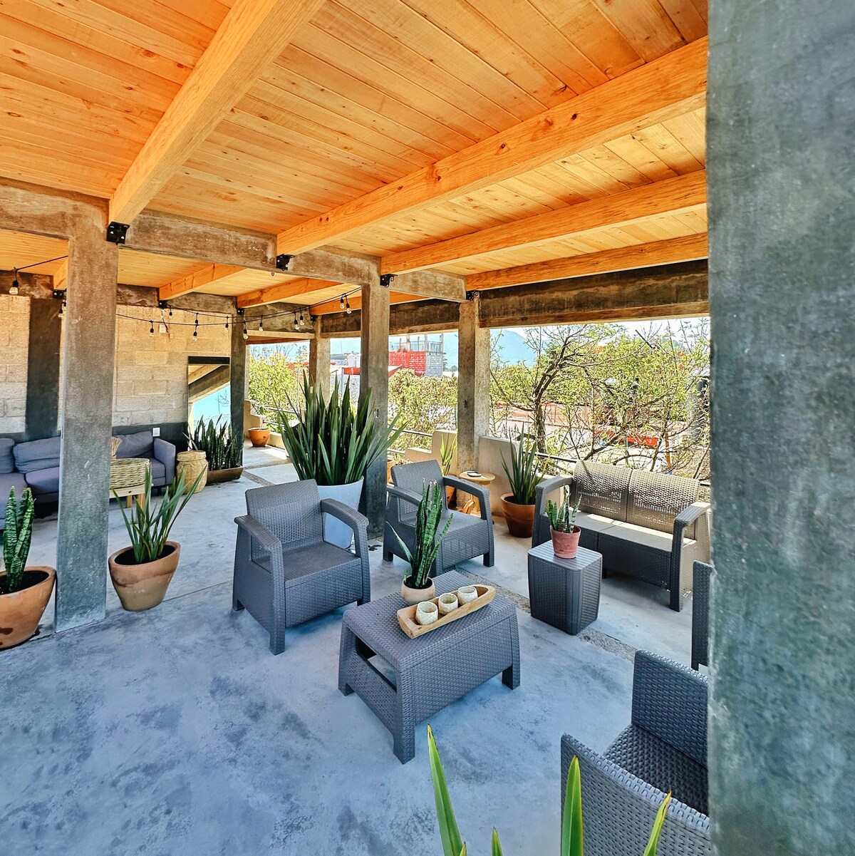 An outdoor terrace is equipped with comfortable seating arrangements, featuring several lounge chairs and tables. Planters with various succulents add greenery to the space. A wood beam ceiling provides a rustic touch, while views of the surrounding landscape are visible in the background.