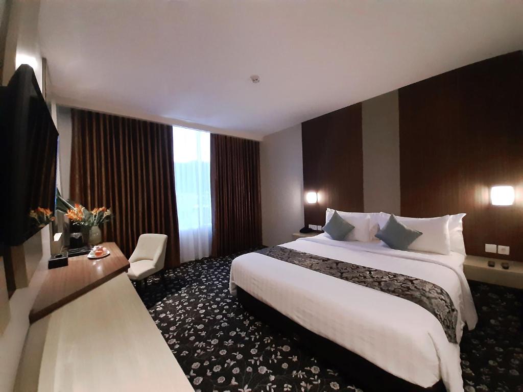 The spacious room features a large double bed adorned with decorative cushions. A flat-screen TV is mounted on the wall opposite the bed, while a small armchair rests near a window dressed with elegant curtains. The carpeted floor is patterned with floral designs.