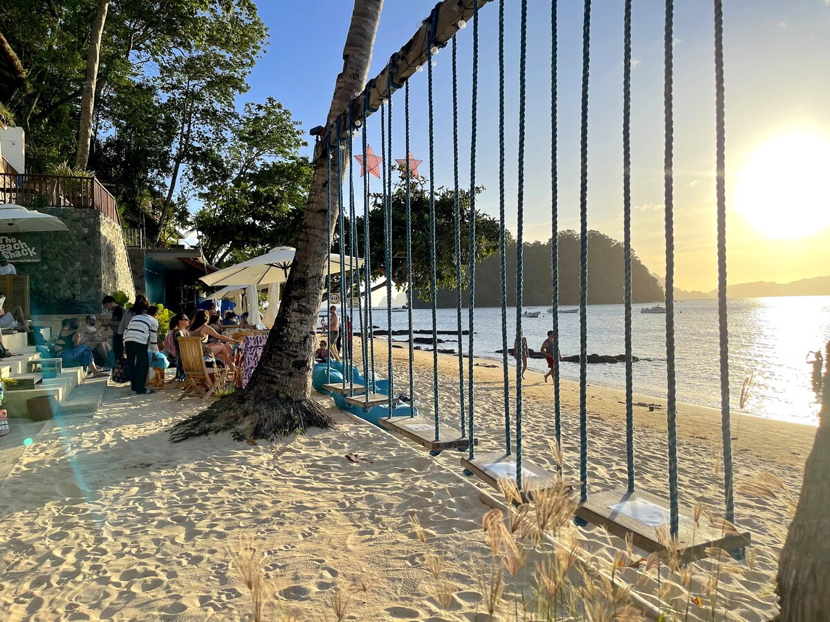 A sandy beach setting is captured, featuring large swings attached to a structural frame. The sun is setting, casting warm orange and yellow hues across the sky. Tables and chairs are visible in the background, with trees providing shade and a serene atmosphere.