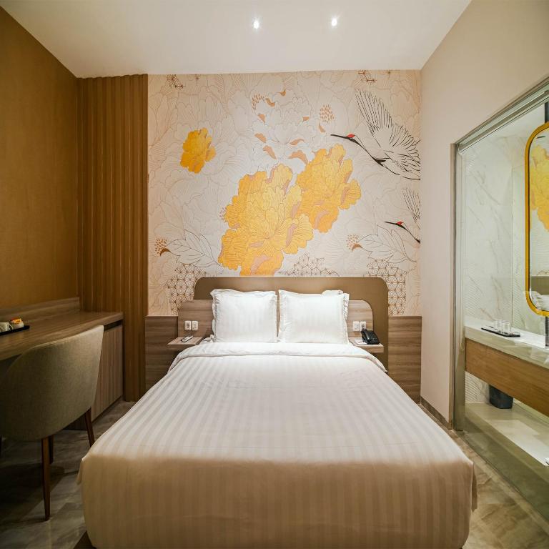 A well-appointed double room features a comfortable bed with crisp linens and accent pillows. A stylish desk and chair are positioned against a textured wall with floral motifs. Natural light fills the space, complemented by soft overhead lighting. A private bathroom is visible through glass doors.