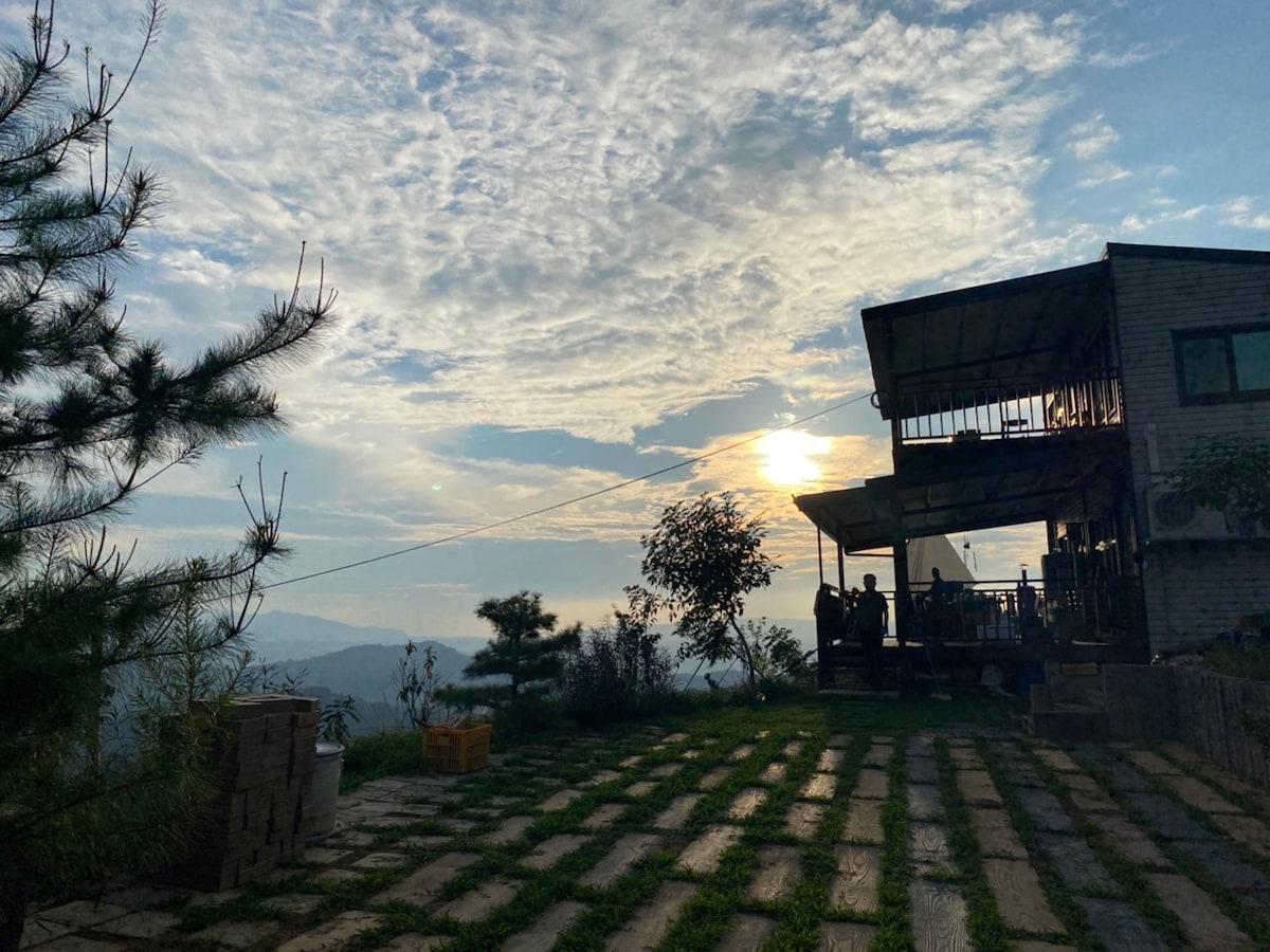 An outdoor view captures the setting sun behind a modern structure. Rolling hills and scattered trees create a natural backdrop. A pathway lined with stone pavers leads towards the building, where guests are observed enjoying the surroundings.