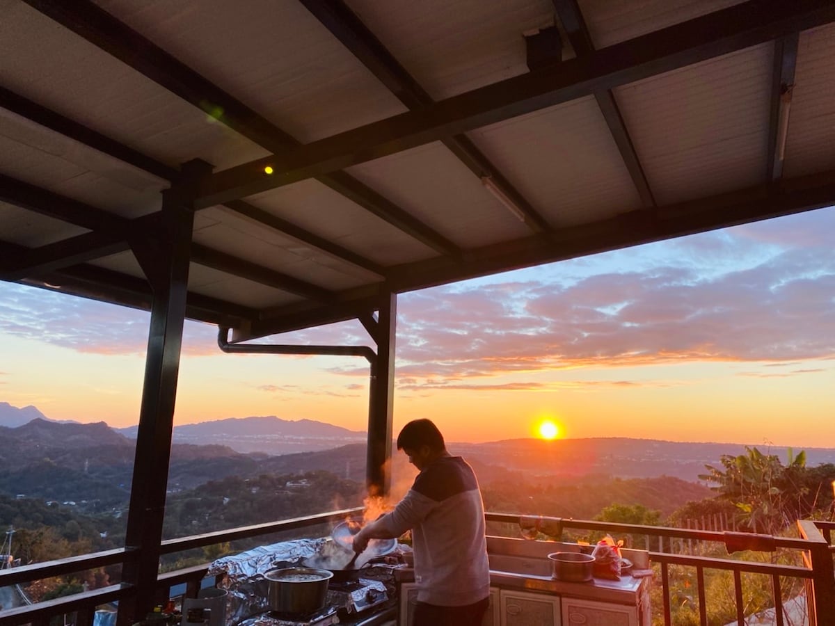 An outdoor kitchen area is set against a stunning sunset, with mountains visible in the background. A person is seen cooking, surrounded by cooking utensils and equipment. The vibrant colors of the sky create a serene atmosphere, enhancing the spacious and open environment.