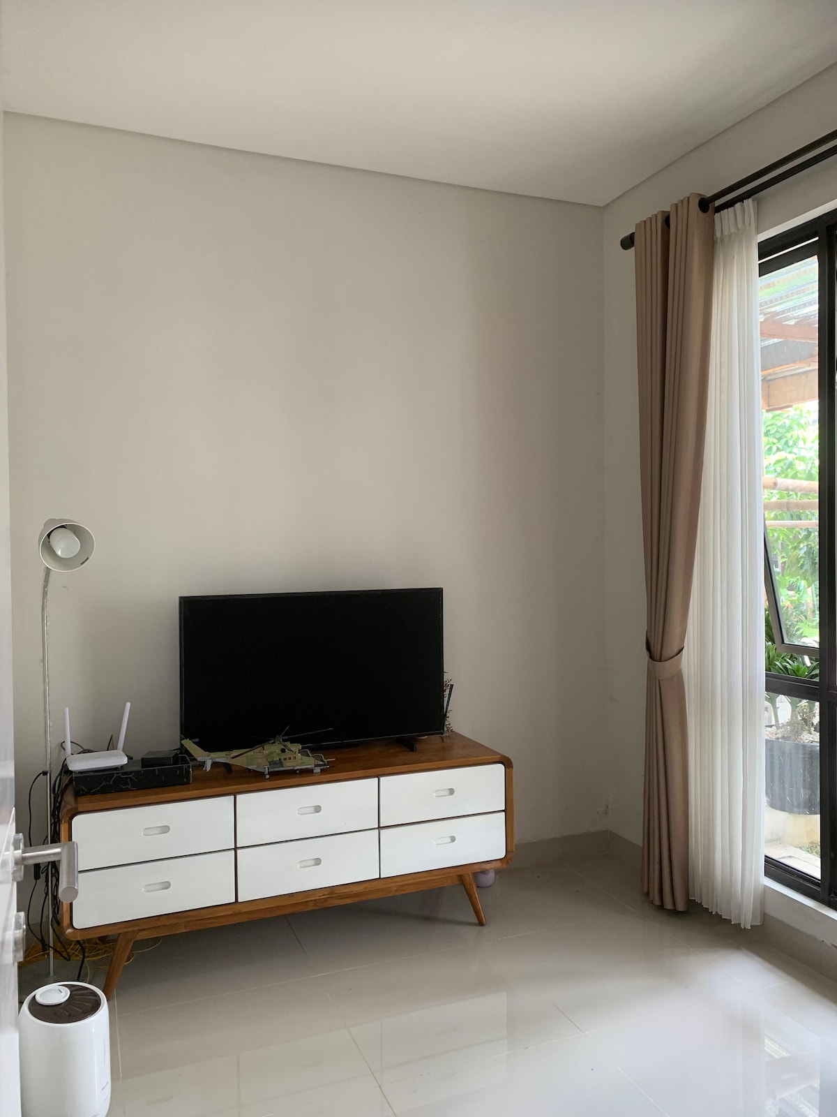 A compact seating area features a wooden sideboard with multiple drawers and a flat-screen television mounted above it. Natural light enters through a large glass window adorned with sheer curtains, enhancing the bright and airy feel of the space.