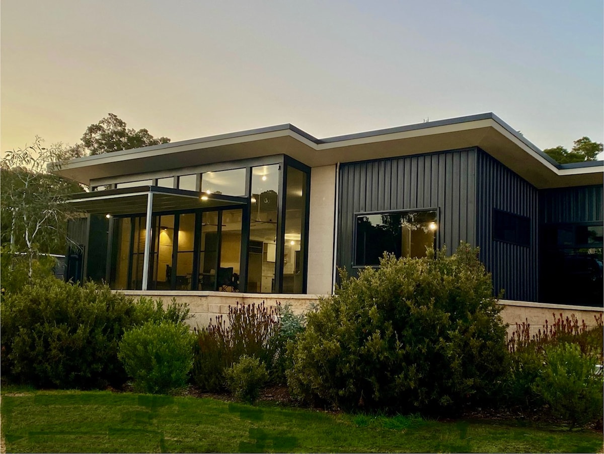 An architecturally designed home exterior is showcased, featuring large glass windows that allow ample natural light. The structure is surrounded by landscaped greenery, with bushes providing a natural border. The setting offers a serene ambiance, highlighted by the evening sky.