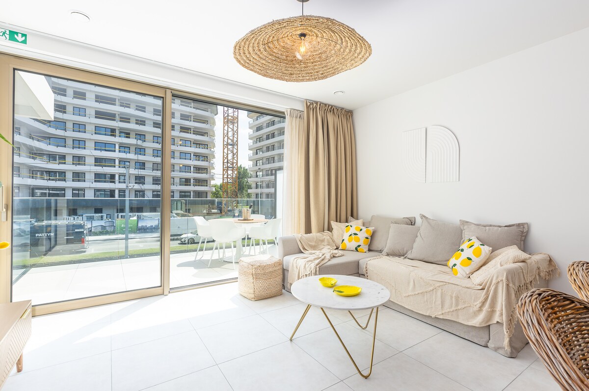 A bright living area is presented with large sliding glass doors leading to a balcony. A light-colored sofa with decorative yellow pillows is accompanied by a round coffee table. Natural light fills the space, enhancing the minimalist design and neutral color palette.
