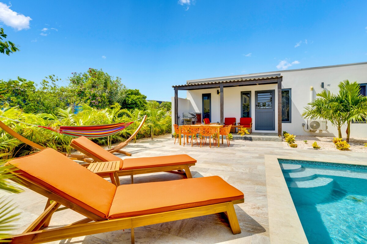 The outdoor area features two orange lounge chairs beside a private pool. A dining table is visible on the patio, surrounded by tropical greenery. A colorful hammock is hung nearby, contributing to the relaxing atmosphere of the villa.