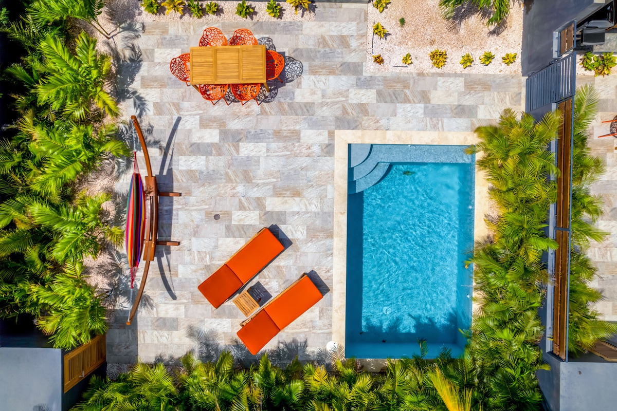 An aerial view captures a private magnesium mineral pool surrounded by lush greenery and stylish lounge chairs. A wooden dining table with colorful seating is visible on the patio, complemented by the pool's clear water and natural stone flooring.