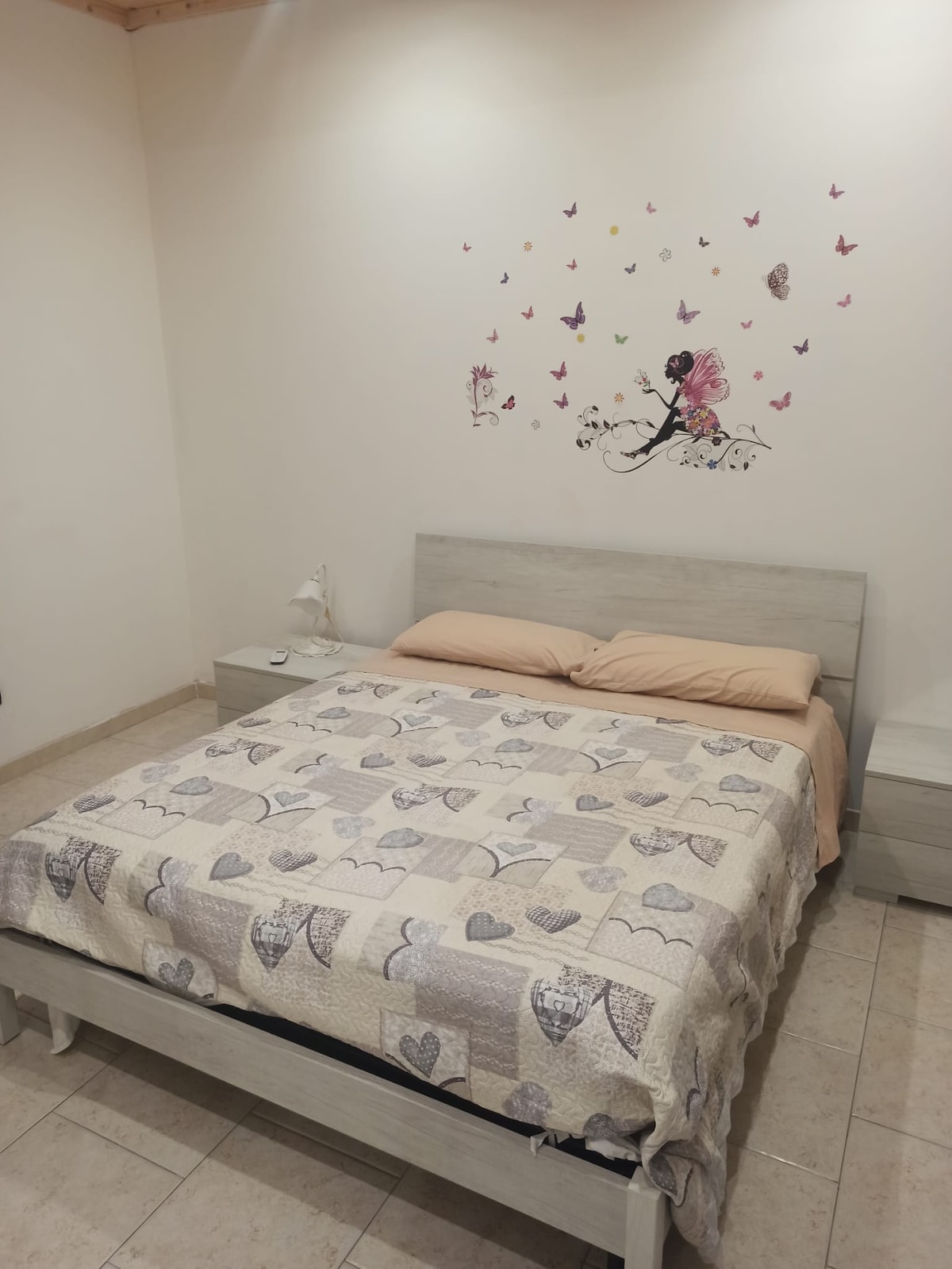 A spacious bed is dressed with a patterned quilt featuring hearts, accompanied by light-colored nightstands on either side. A delicate wall decoration of butterflies adds a subtle touch to the neutral-colored walls, creating a serene sleeping environment.