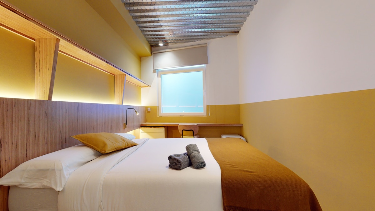 A cozy bedroom is presented with a spacious bed dressed in white linens and a brown throw. Soft lighting highlights the wooden accents and a desk is positioned next to the bed. A large window allows natural light to illuminate the room.