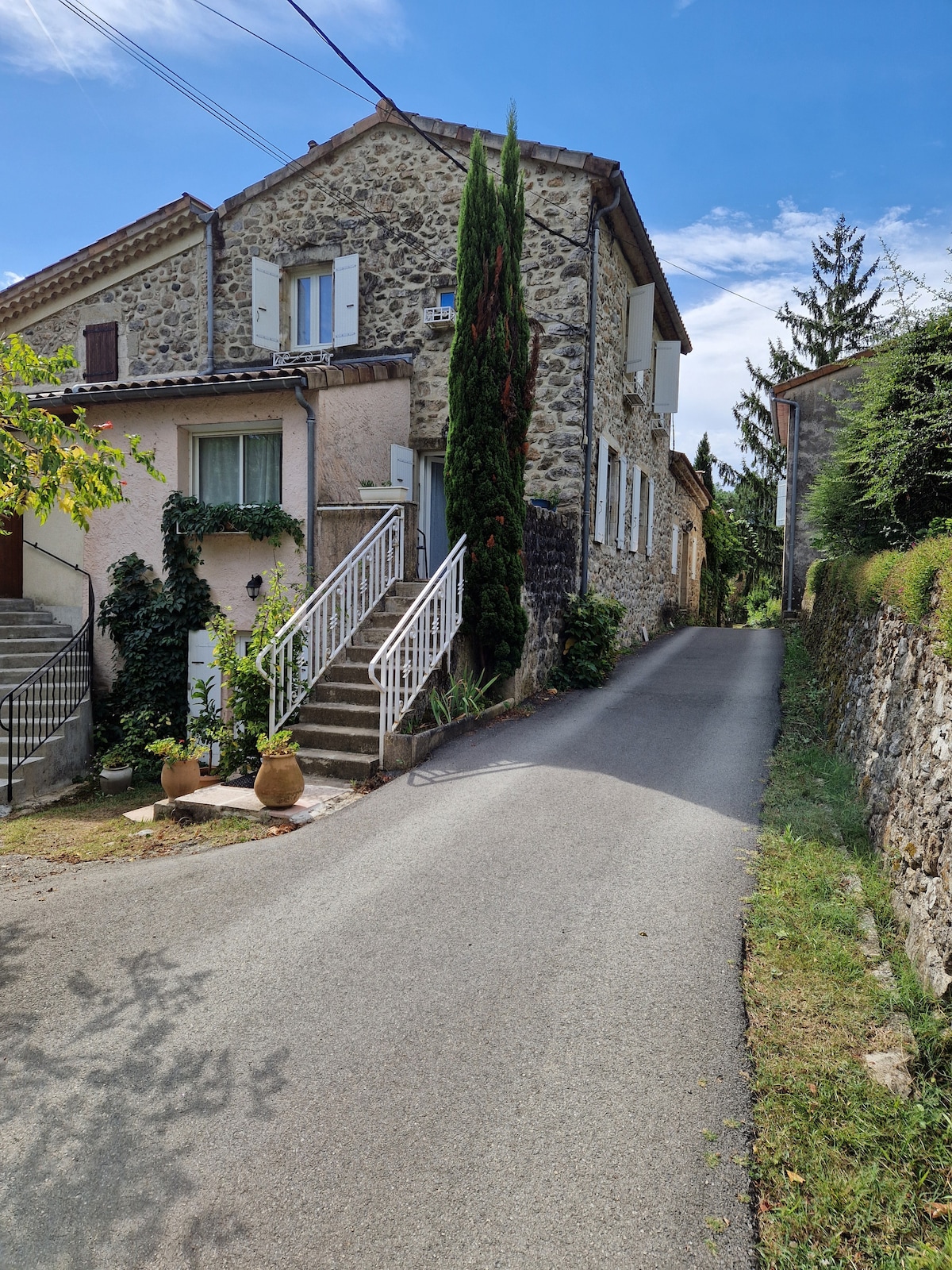 A peaceful stone house is positioned on a gently sloping street, surrounded by greenery. The exterior features a combination of smooth and textured stone surfaces, with white shutters adorning the windows. Planters with foliage add a touch of greenery along the entrance stairs.