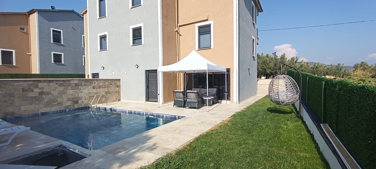 The exterior of the villa features a private pool surrounded by a stone patio. A shaded seating area with a tent offers comfortable furnishings. The lush green lawn complements the neutral exterior of the two-story building, which has multiple windows allowing for ample natural light.