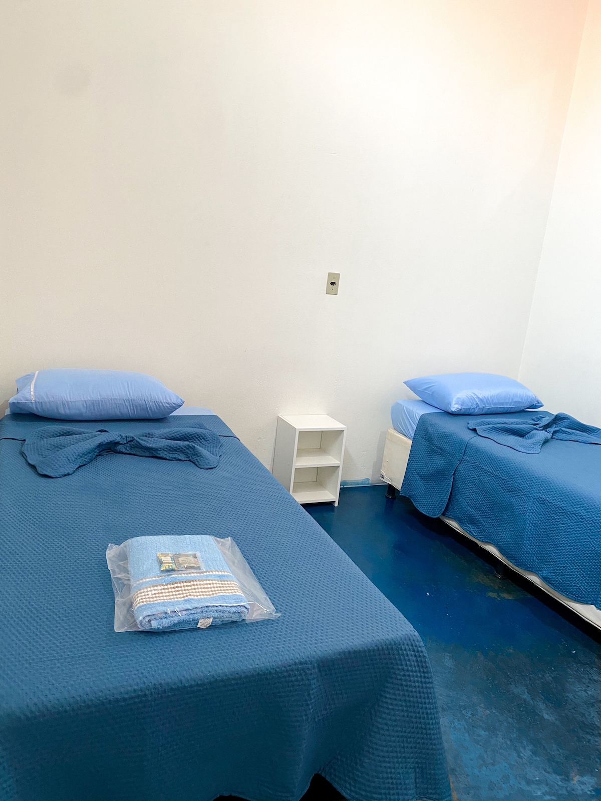 The image features a comfortable sleeping area with two single beds, each dressed in light blue linens. A small bedside table is positioned between the beds, while clean towels are neatly arranged on one bed. The walls are painted white, creating a bright and spacious atmosphere.