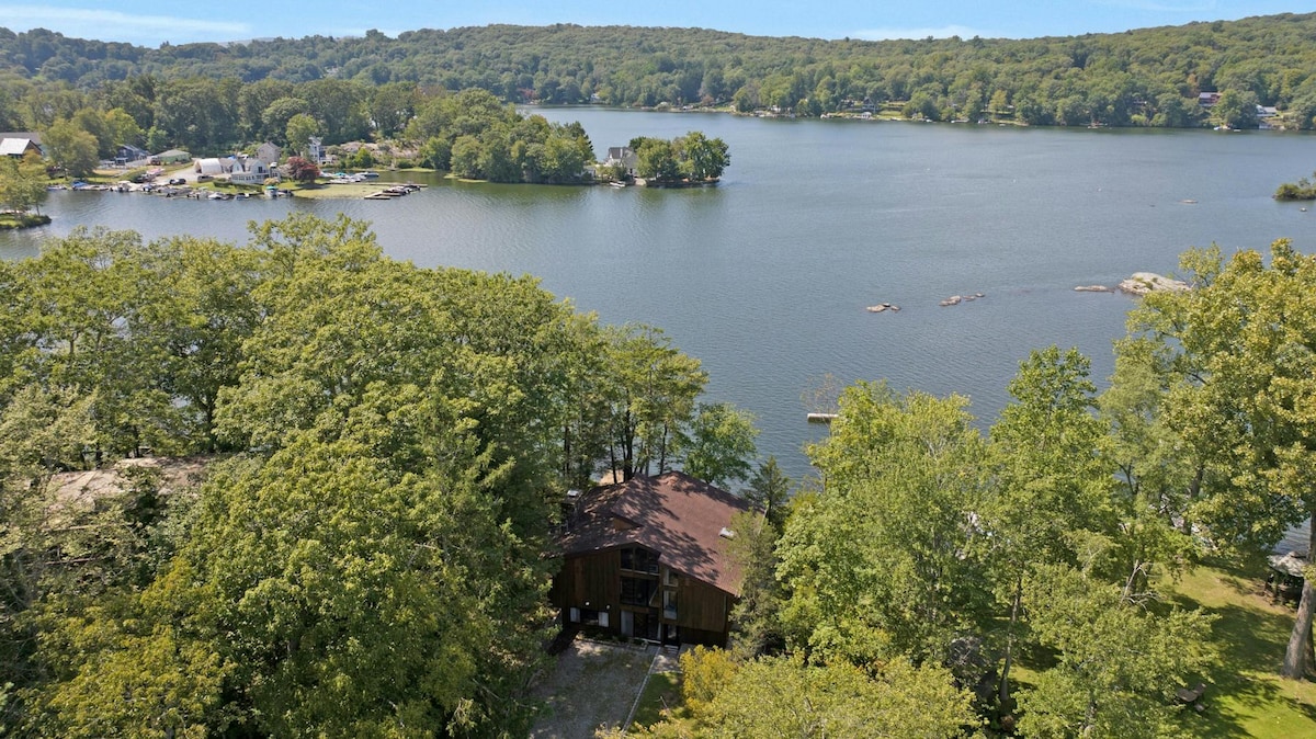 An aerial view captures the peaceful lakefront home nestled among lush trees. The sparkling water stretches out towards the horizon, reflecting the surrounding greenery. The property offers access to the lake, enhancing the serene outdoor setting.