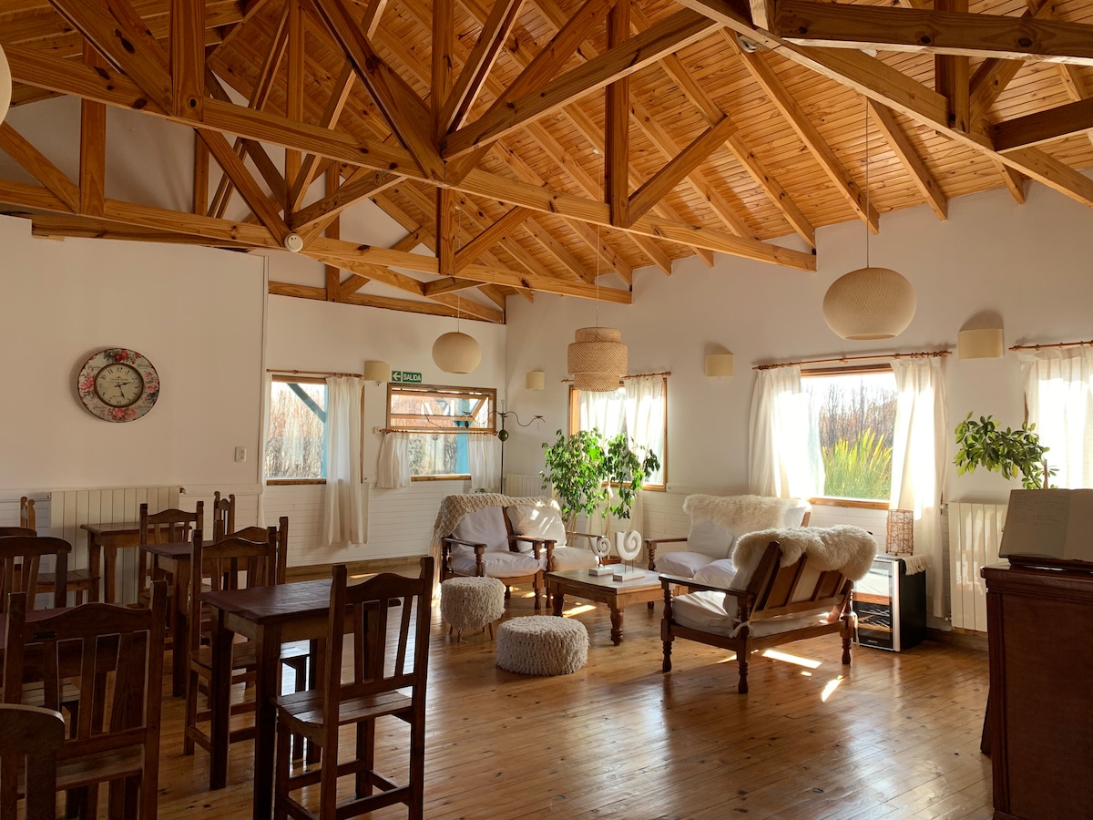 An open living area is highlighted by high wooden beams and natural light. Comfortable seating options are complemented by greenery, while neutral tones create a calming ambiance. Large windows allow views of the surrounding landscape, and various light fixtures provide a warm glow.