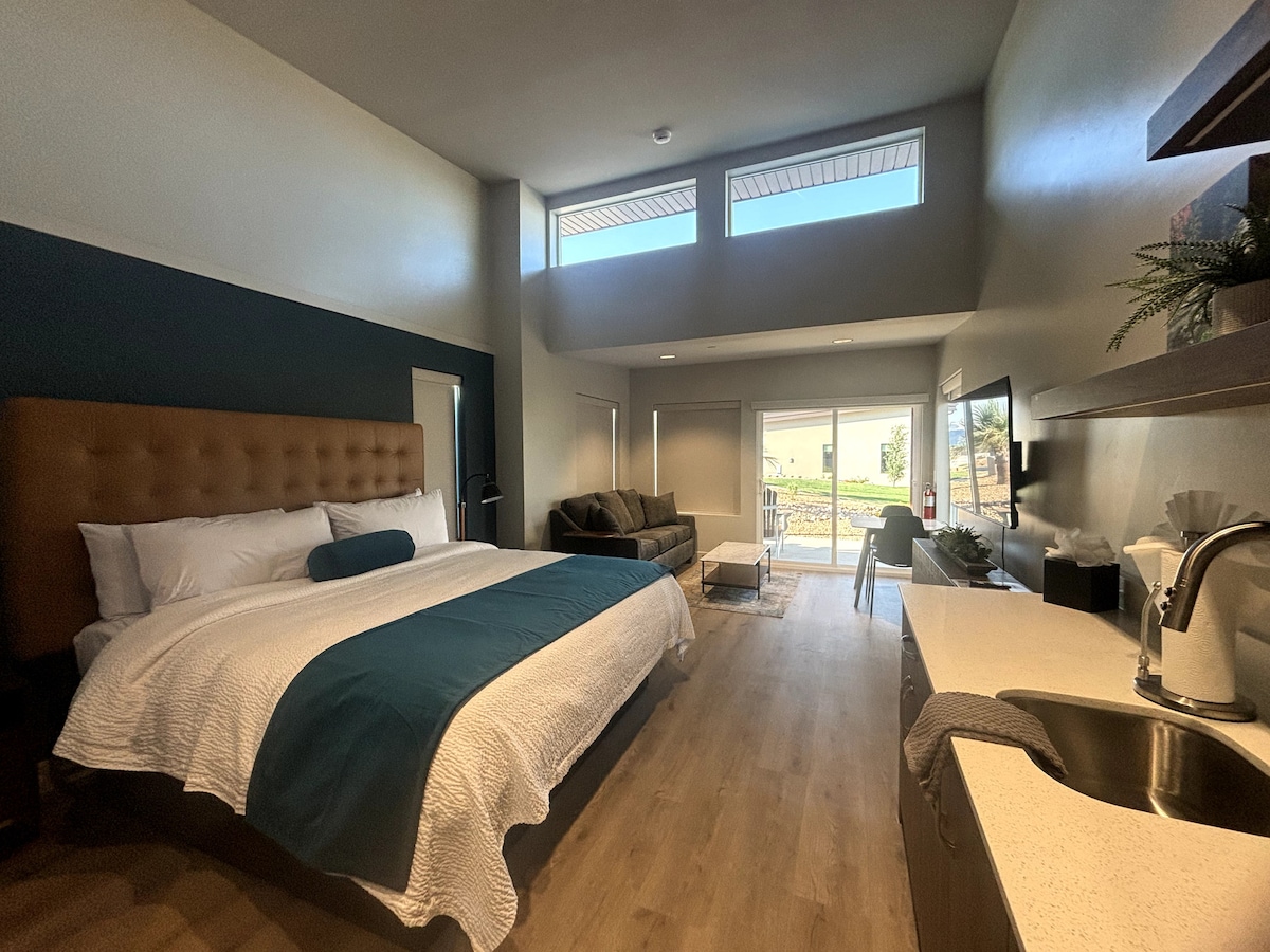 A spacious room features a king bed with a modern headboard and crisp linens. A cozy sofa is positioned nearby, and large windows allow natural light to illuminate the space. A small kitchenette area is visible, complete with a sink and coffee maker.