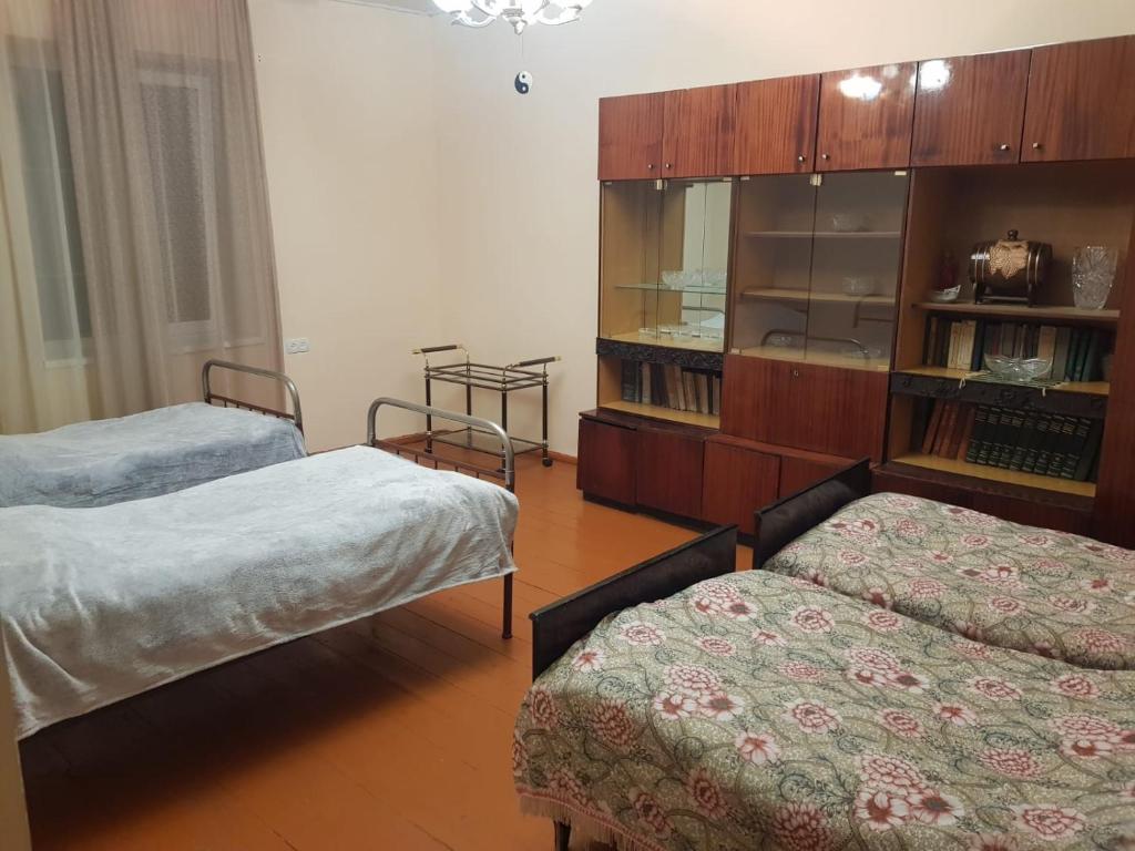 The room features two single beds, each covered with soft blankets. A spacious wooden cabinet with glass doors is present, displaying various items. Light drapes adorn the window, allowing natural light to brighten the room's simple and functional design.