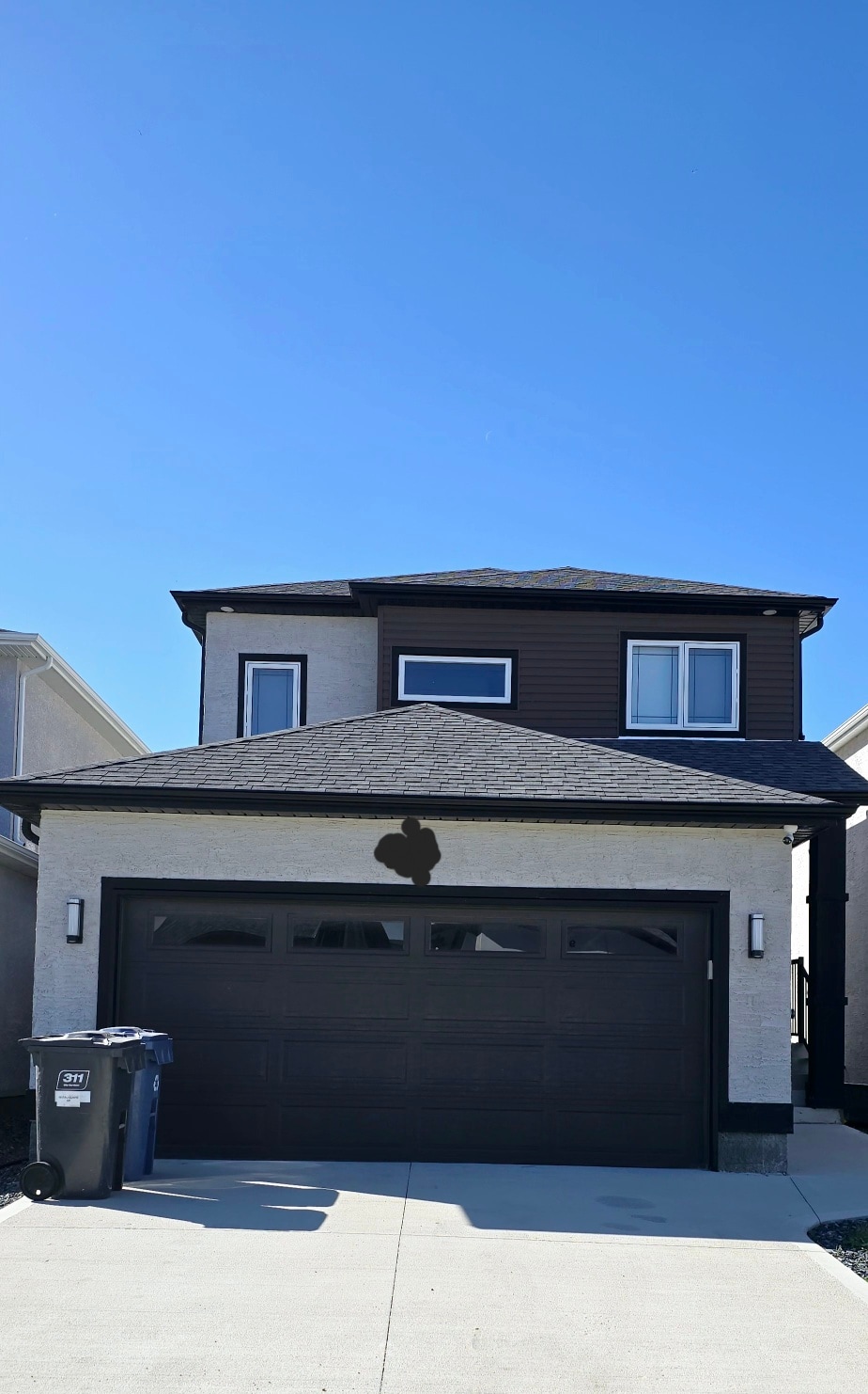 A two-story house features a combination of light-colored stucco and dark wood accents. A spacious two-car garage is visible, complemented by driveway space. The clear blue sky above enhances the home's inviting exterior appearance.